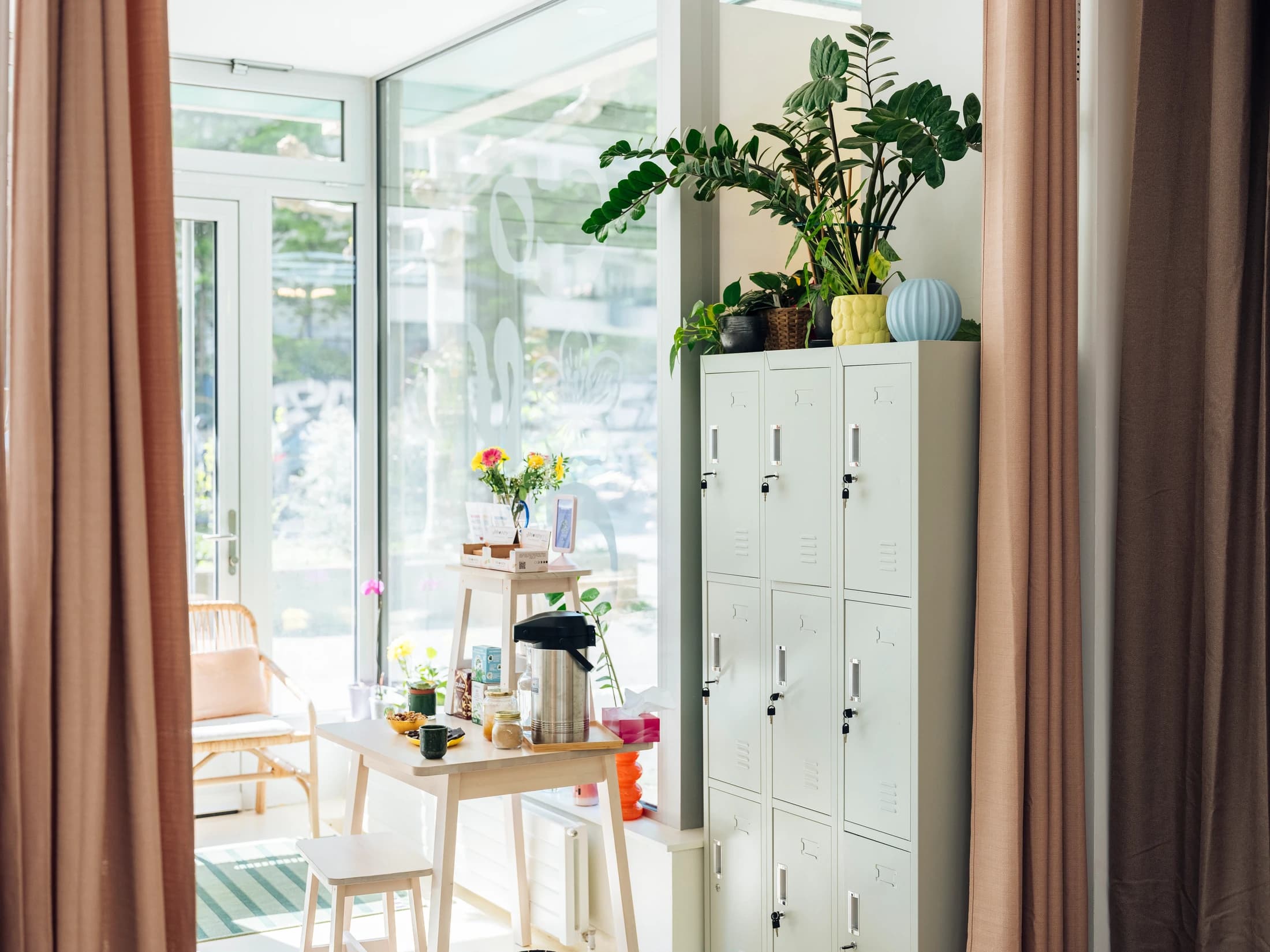 Coin accueil COMOCO — banquette en rotin, plantes vertes luxuriantes, fleurs fraîches, lumière du jour entrant par la baie vitrée