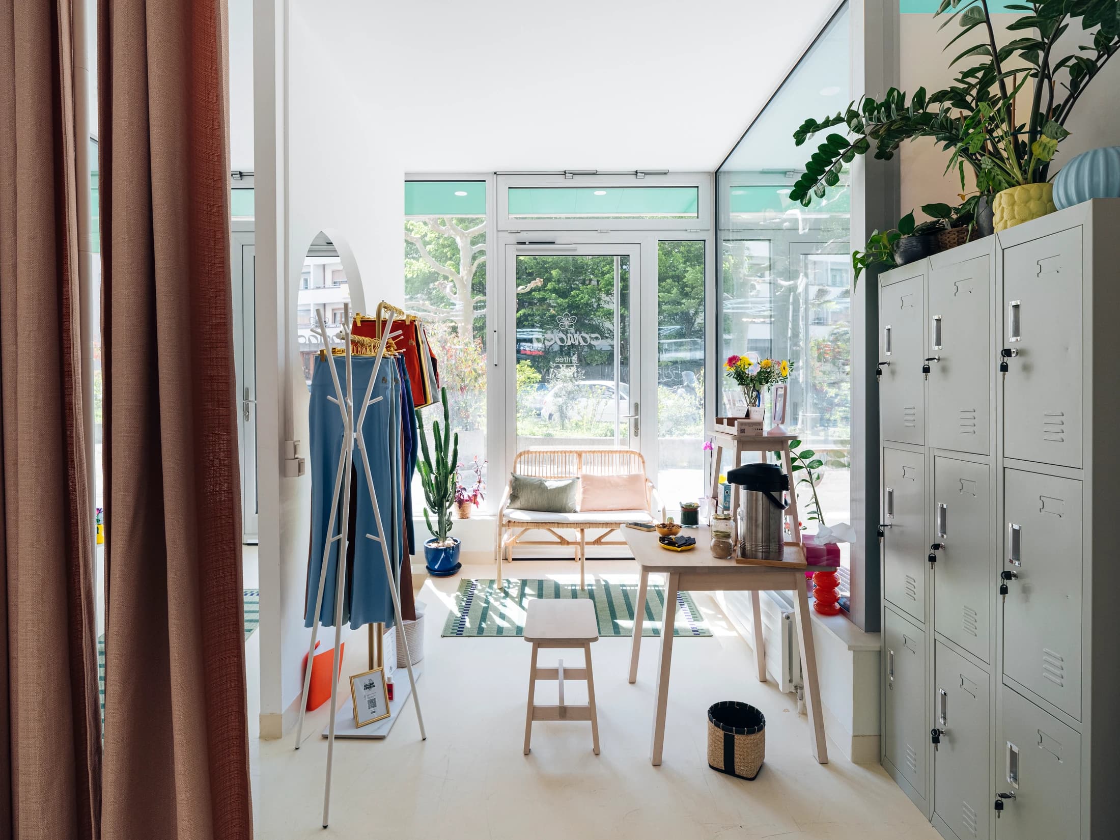 Coin boutique — portant de vêtements colorés, banquette osier, plantes vertes, atmosphère lumineuse
