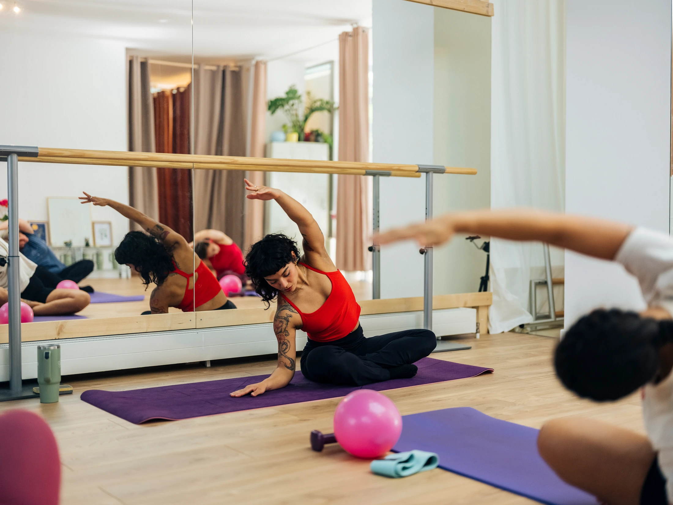Cours de pilates en action : élève en débardeur rouge en étirement latéral devant les barres et miroirs, tapis violets, balle rose