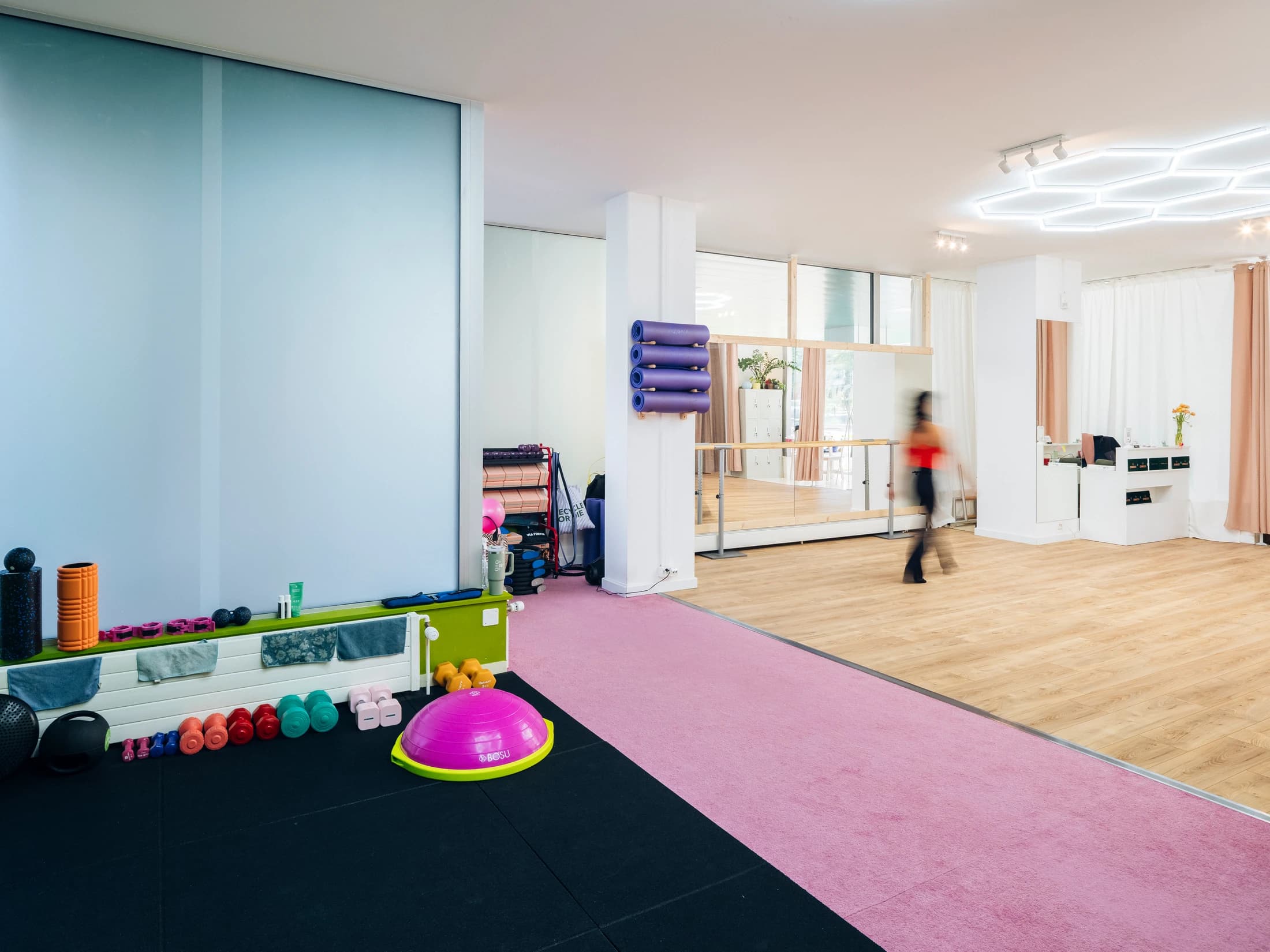 Petite salle de sport — haltères colorés, ballon BOSU rose, mousses, moquette rose et noire, intervenante en mouvement