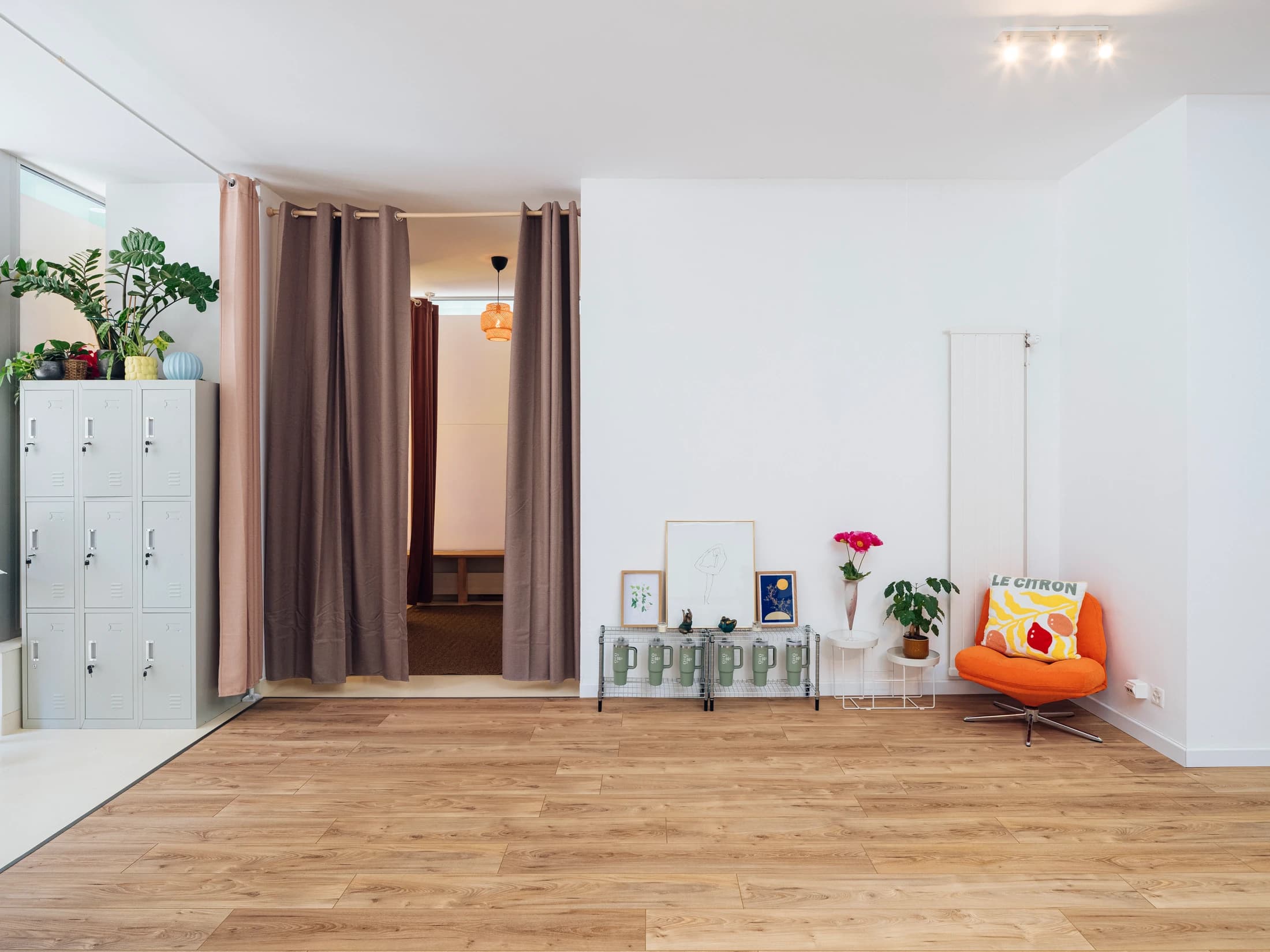 Vestiaire COMOCO — lockers verts pâles, rideaux taupe doublés rose, fauteuil orange avec coussin Le Citron, cadres artistiques